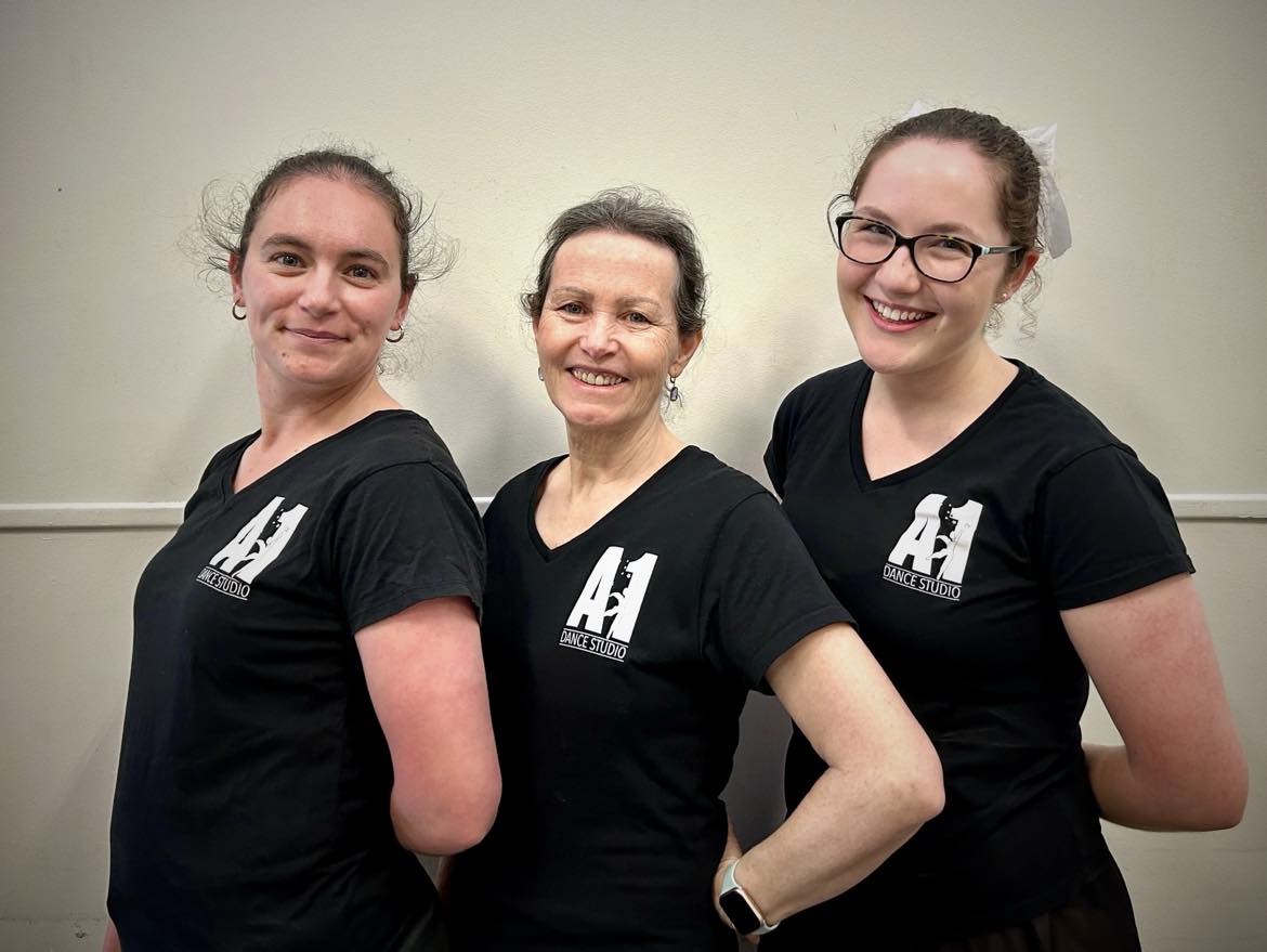 Three smiling women wearing matching black t-shirts emblazoned with white A1 dance logos, each with their hands behind their back and standing at a 45 degree angle to the camera.